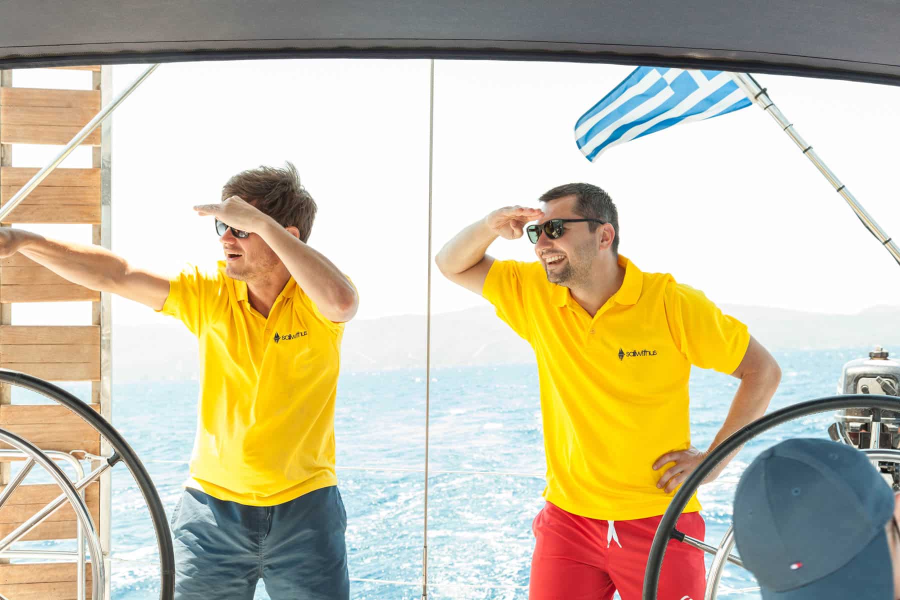 Zwei Männer in gelben Poloshirts und Sonnenbrillen auf einer Segelyacht, die ihre Augen abschirmen, um in die Ferne zu blicken, während im Hintergrund eine griechische Flagge weht.