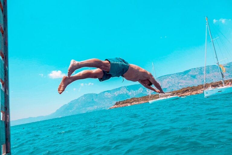 Eine Person macht einen Rückwärtssalto von einem Boot ins Meer, über ihr ein klarer blauer Himmel und in der Ferne Berge. In der Nähe sind zwei Segelboote zu sehen, ideal für Segelbegeisterte.