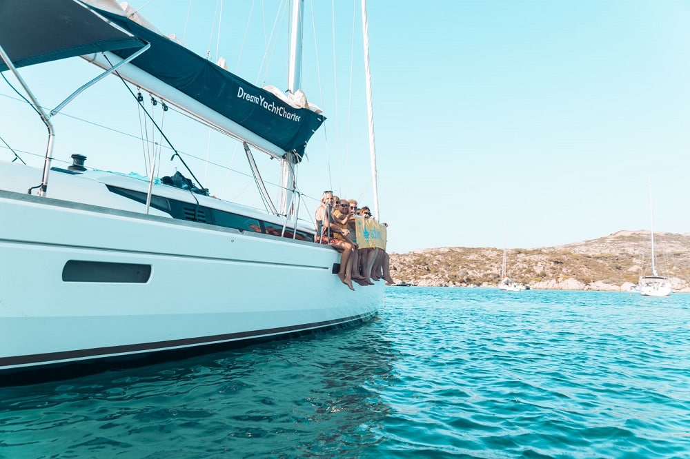 Eine Gruppe von Menschen lächelt und posiert während ihres Segelurlaubs für ein Foto auf dem Bug eines Segelboots, das im klaren, türkisfarbenen Wasser vor Anker liegt, mit felsigen Hügeln im Hintergrund.