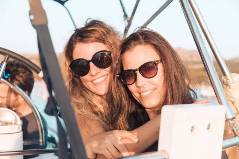 Zwei Frauen mit Sonnenbrillen lächeln und machen an einem sonnigen Tag ein Selfie auf einem Segeltörn mit offenem Verdeck. Auf ihren Gesichtern ist deutlich Freude und Kameradschaft zu erkennen.