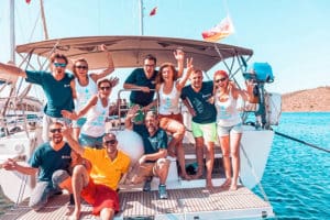Eine Gruppe von neun Erwachsenen posiert fröhlich auf dem Deck einer Segelyacht, winkt und lächelt unter einem klaren blauen Himmel, umgeben von ruhigem blauen Wasser.