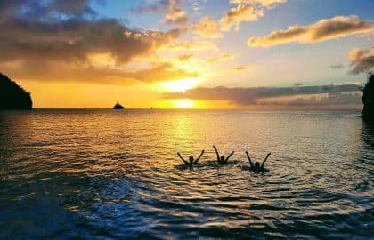 Ein leuchtender Sonnenuntergang über einem ruhigen Meer mit Silhouetten von Menschen, die freudig ihre Hände über das Wasser strecken, eingerahmt von Klippen auf beiden Seiten. Ein entferntes Segelboot, bereit für ein Segelurl