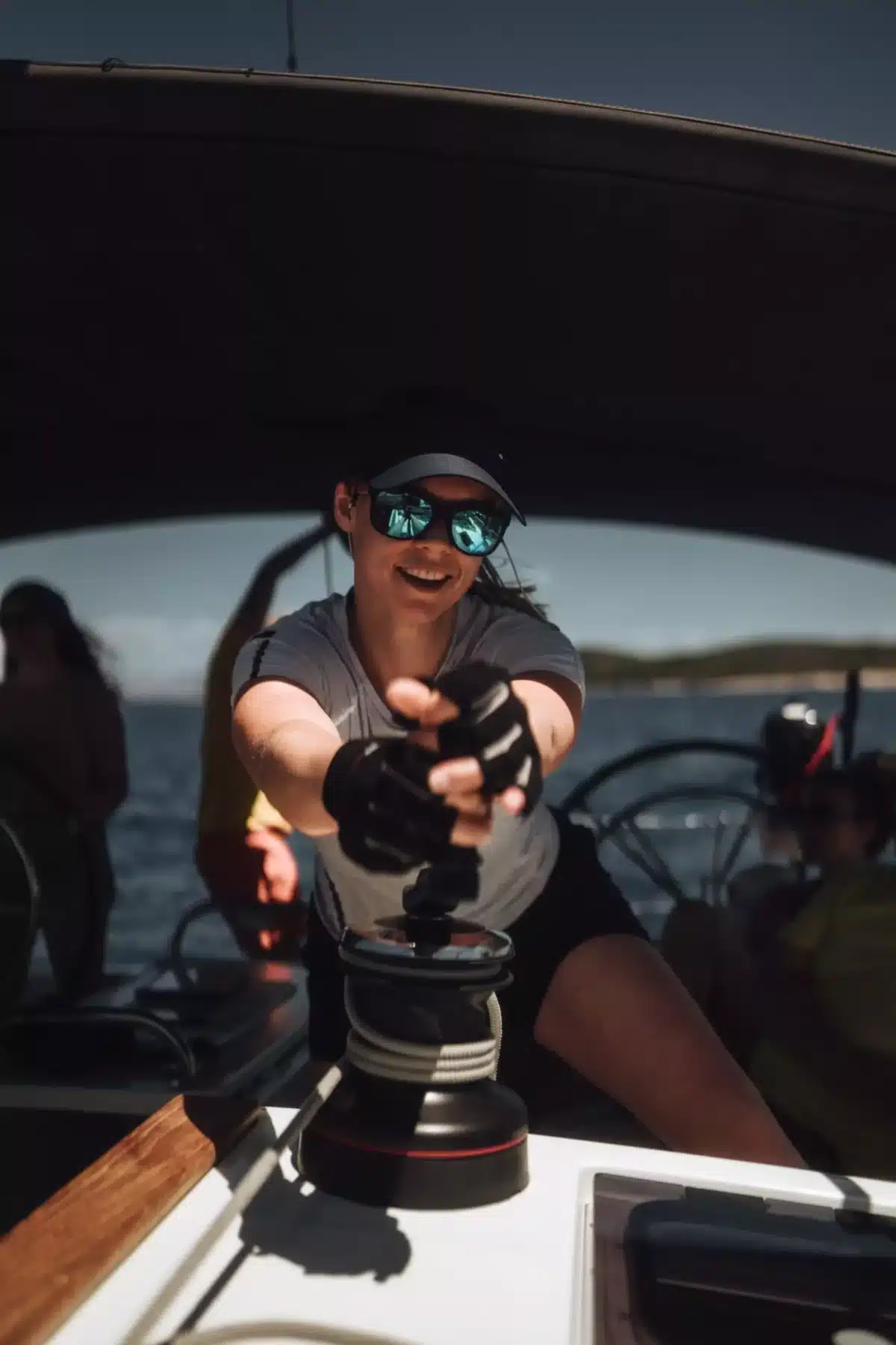 Eine Frau mit Sonnenbrille und Mütze steuert eine Segelyacht mit einer Winschkurbel und lächelt an einem sonnigen Tag unter einem Schattendach.
