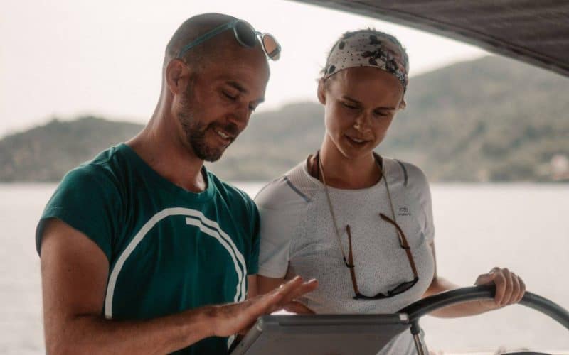 Ein Mann und eine Frau sind auf einem Boot und besprechen wahrscheinlich Navigationspläne für ihren Segelurlaub. Der Mann deutet auf eine Karte oder einen Bildschirm, beide tragen Freizeitkleidung, mit dem Meer und
