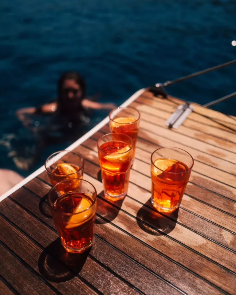 Vier Gläser Eistee mit Zitronenscheiben auf dem Holzdeck einer Segelyacht, mit dem klaren blauen Ozean im Hintergrund und einer Person, die neben der Yacht schwimmt.