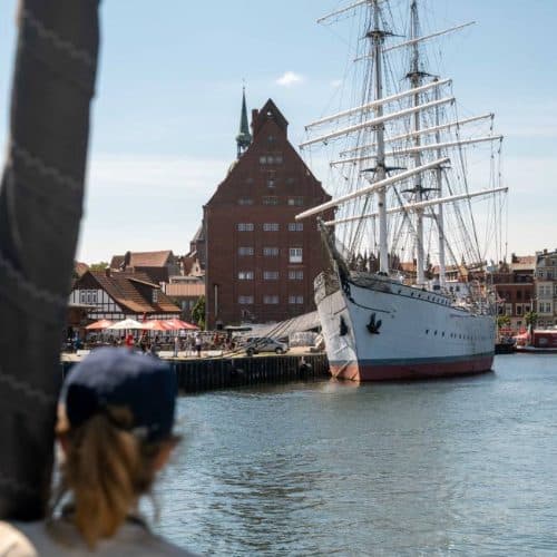 Eine Person mit einer Mütze beobachtet eine große weiße Segelyacht, die an einer sonnigen Uferpromenade vor Anker liegt. Im Hintergrund sind ein historisches rotes Gebäude und andere Strukturen zu sehen.