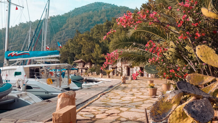 Ein ruhiger Yachthafen mit Segelyachten, die neben einem gepflasterten Gehweg vor Anker liegen, gesäumt von üppigem Grün und leuchtend rosa blühenden Sträuchern, mit Hügeln im Hintergrund.