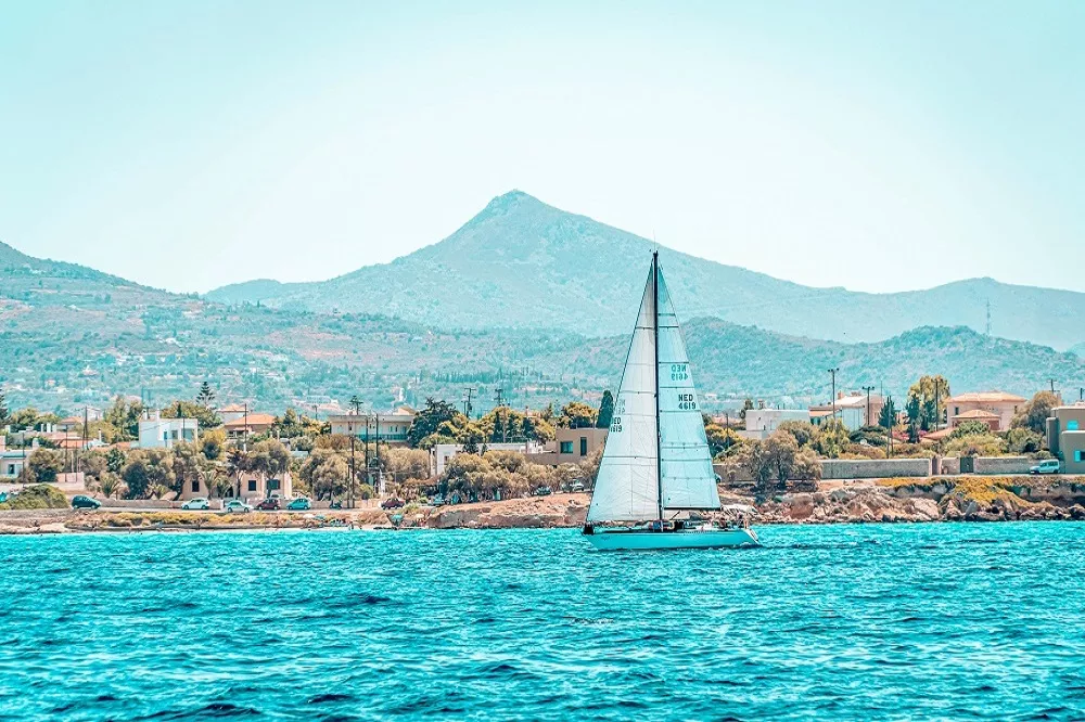 Eine Segelyacht mit einem weißen Segel auf einem leuchtend blauen Meer vor einer Küstenstadt mit verstreuten Häusern und einem Berg im Hintergrund unter einem klaren Himmel.