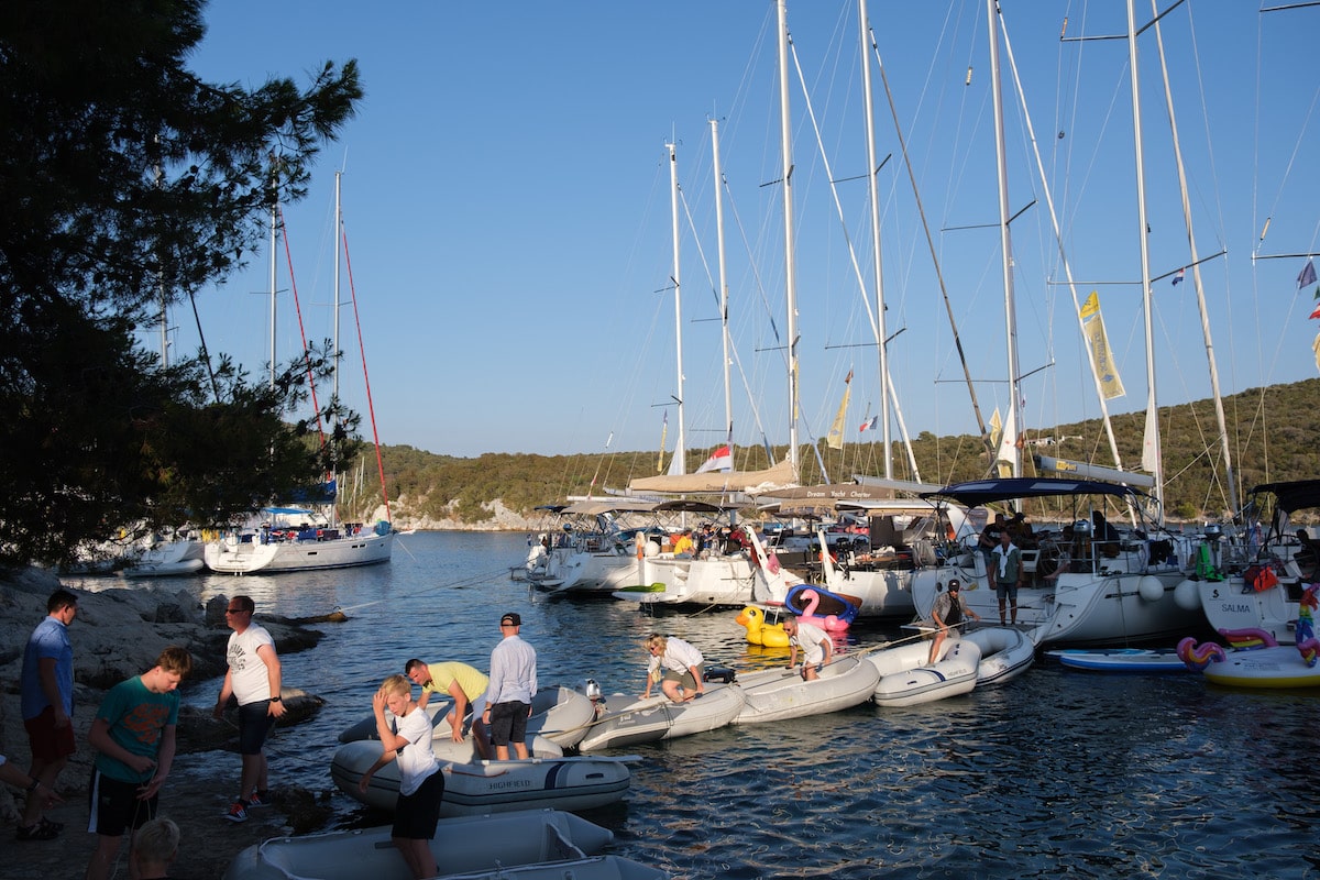 Eine belebte Bucht mit zahlreichen Booten und Yachten, die dicht beieinander liegen, Menschen, die sich am Ufer und in kleinen Schlauchbooten tummeln, umgeben von üppigem Grün unter einem klaren Himmel, perfekt für