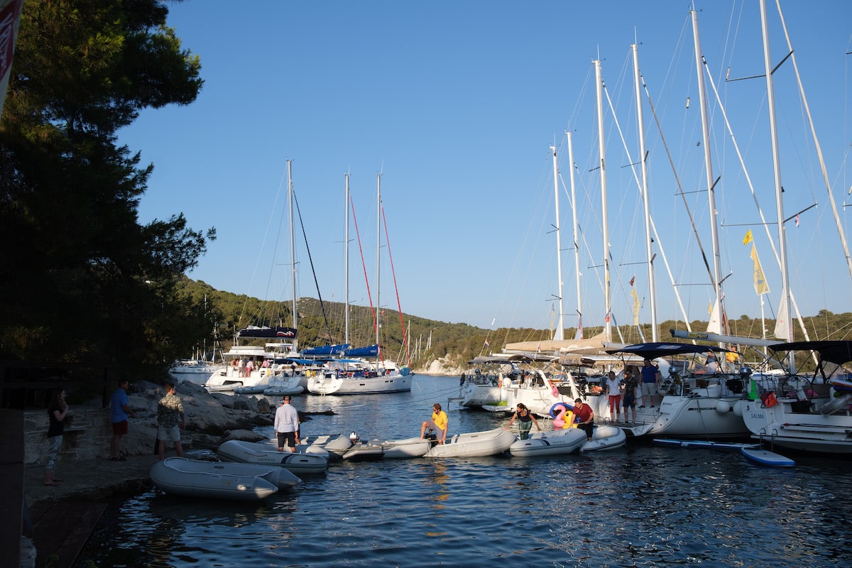 Segelyachten liegen in einer ruhigen, von Bäumen gesäumten Bucht vor Anker. An einem sonnigen Tag tummeln sich am Ufer und in kleinen Schlauchbooten Menschen.