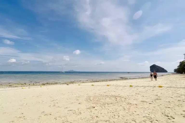 Zwei Menschen gehen an einem Sandstrand spazieren, der Himmel ist klar, das Meer ruhig und blau, in der Ferne ist eine Segelyacht zu sehen. Sonnenlicht erhellt die Szene.