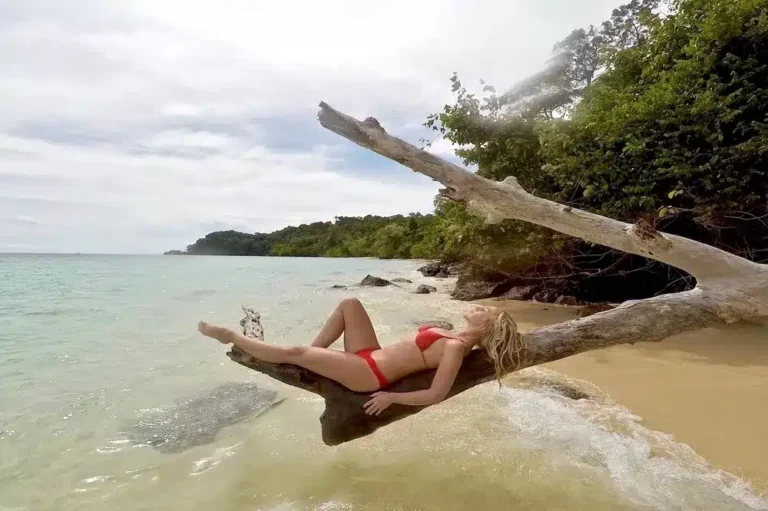 Eine Frau in einem roten Badeanzug entspannt sich auf einem großen Treibholzstamm an einem Sandstrand, mit üppigem Grün und einem ruhigen Meer im Hintergrund, in der Nähe der Stelle, an der Segelyachten festgemacht sind