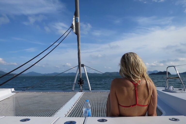 Eine Frau mit blondem Haar sitzt auf einer Segelyacht und blickt auf eine malerische Aussicht auf ferne Berge und einen wolkenverhangenen Himmel über einer großen Wasserfläche.