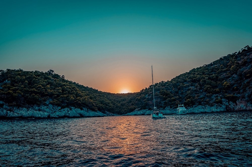 Sonnenuntergang über einer ruhigen Bucht mit der Silhouette einer Segelyacht zwischen bewaldeten Hügeln, die das orangefarbene Leuchten auf dem Wasser reflektiert.