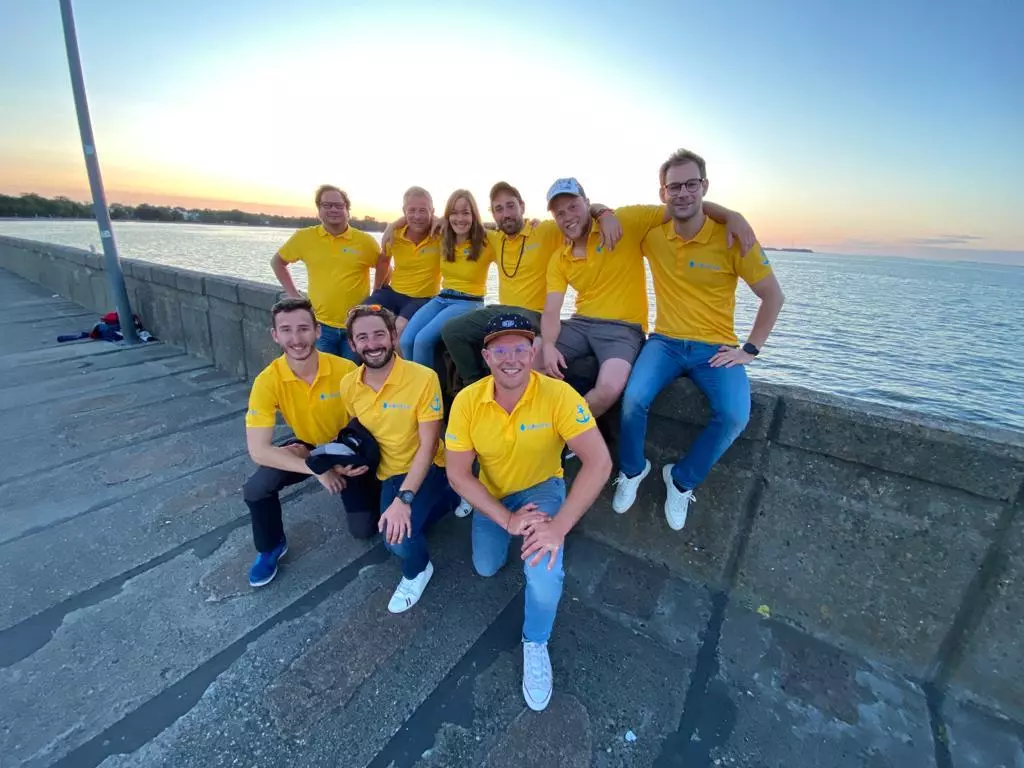 Eine Gruppe von acht Personen in passenden gelben Hemden lächelt und posiert während eines Segelurlaub-Sonnenuntergangs auf Steinstufen neben einem Gewässer.