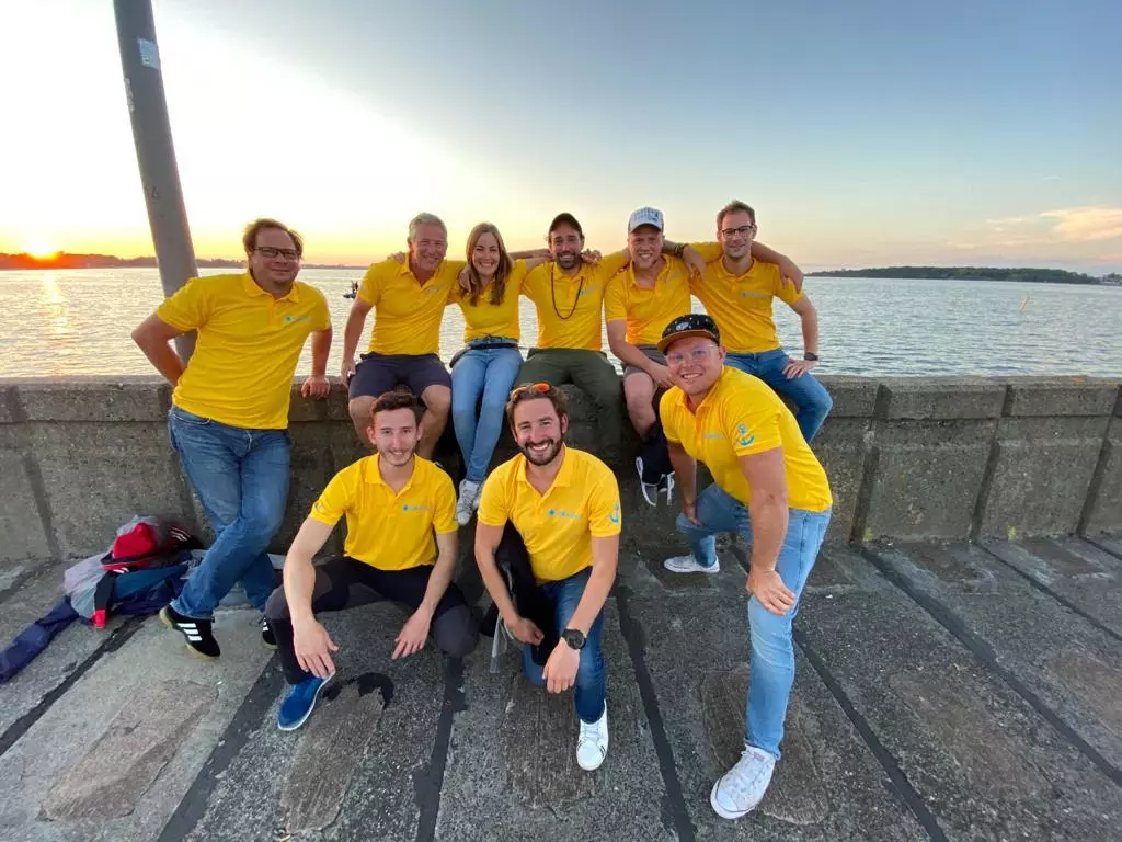 Neun Menschen in gelben Hemden, die lächelnd gemeinsam bei Sonnenuntergang auf einer Segelyacht posieren.