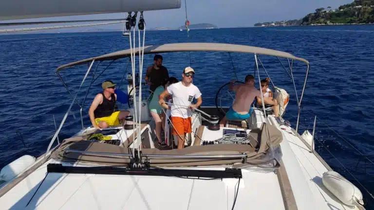 Eine Gruppe von Menschen genießt einen sonnigen Tag auf einem Katamaran. Ein Mann steht und lächelt in die Kamera, andere sitzen oder stehen um ihn herum. Im Hintergrund ist die Küste zu sehen.