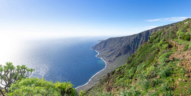 Ein Panoramablick auf eine Küste zeigt einen steilen, grün bewachsenen Berghang mit Blick auf das Meer. Das klare blaue Wasser trifft auf das zerklüftete Ufer, wölbt sich nach innen und erstreckt sich unter einem hellen, klaren Himmel in die Ferne. Im Vordergrund ist spärliche Vegetation zu sehen – perfekt für ein Segelurlaub-Abenteuer.