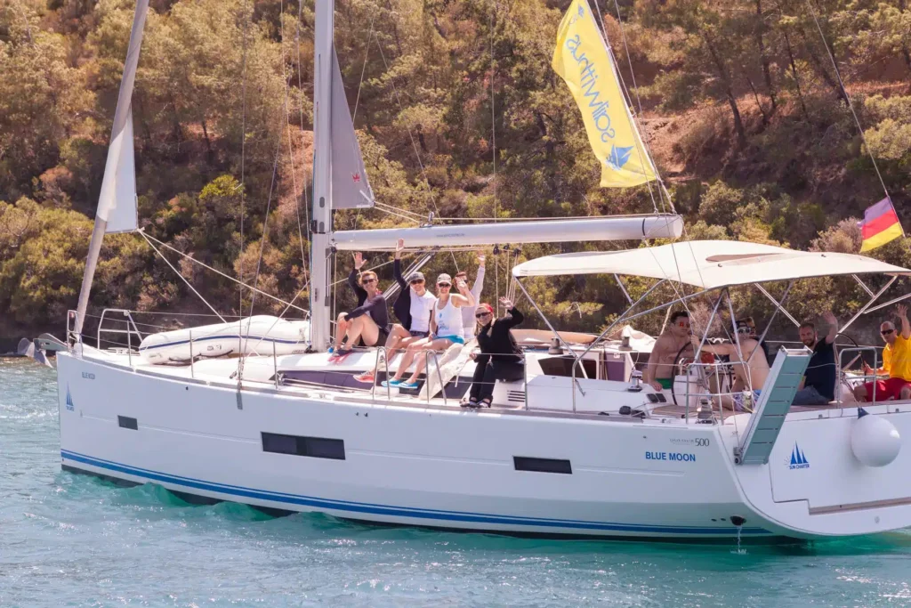 Eine Gruppe von Menschen genießt einen sonnigen Tag auf einem großen weißen Segelboot namens „Blue Moon“. Sie winken und lächeln in die Kamera, wahrscheinlich während ihres Skippertrainings. Das Segelboot liegt auf einem ruhigen Gewässer, im Hintergrund ist eine bewaldete Küste zu sehen. Am Mast des Bootes wehen Flaggen. Mitsegeln vs. Kreuzfahrt: Welche Reiseart passt zu Dir?