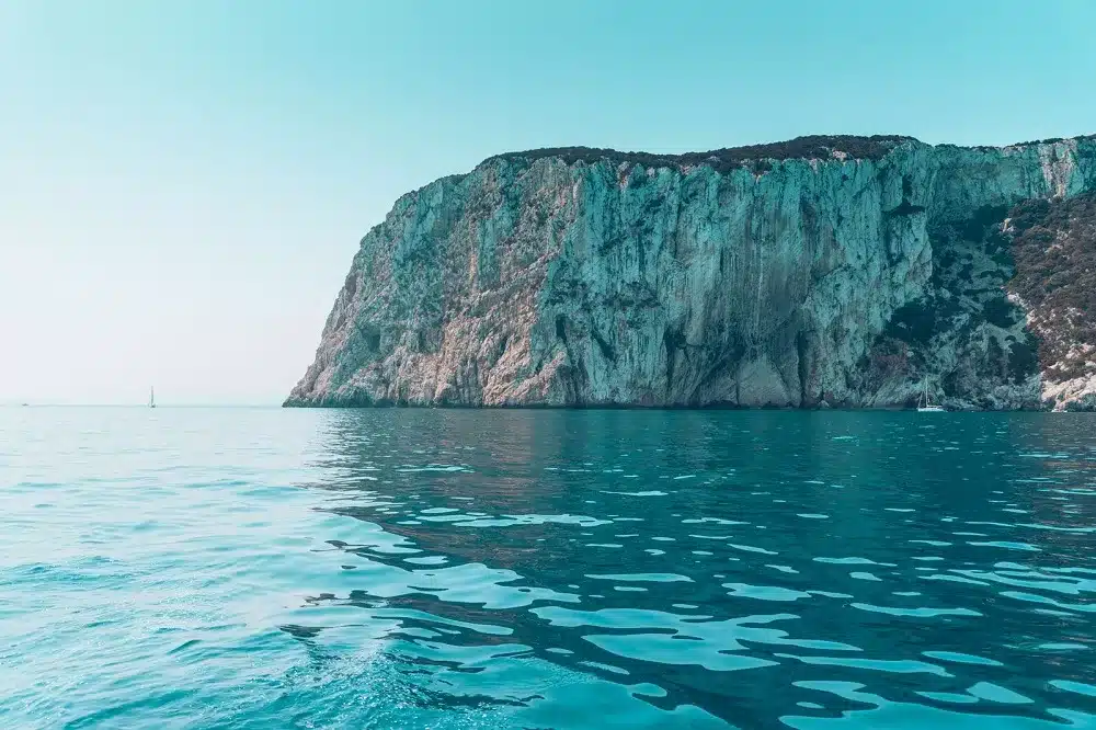 Eine ruhige Meereslandschaft mit einer hoch aufragenden, zerklüfteten Klippe mit grünen Flecken über dem ruhigen, türkisfarbenen Wasser und einer Segelyacht unter einem klaren Himmel.