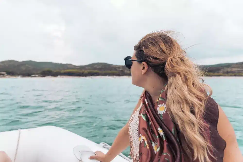 Eine Frau mit langem, welligem Haar und Sonnenbrille blickt von der Seite einer Segelyacht aus, in einen geblümten Schal gehüllt, auf eine Küstenlandschaft.