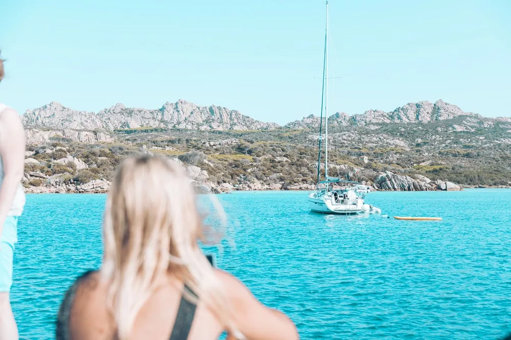 Ein Segelboot, das im kristallklaren, türkisfarbenen Wasser nahe einer felsigen Küste vor Anker liegt. Im Vordergrund sind die verschwommenen Gestalten zweier Menschen zu sehen, die die malerische Aussicht auf den Segelurlaub genießen.