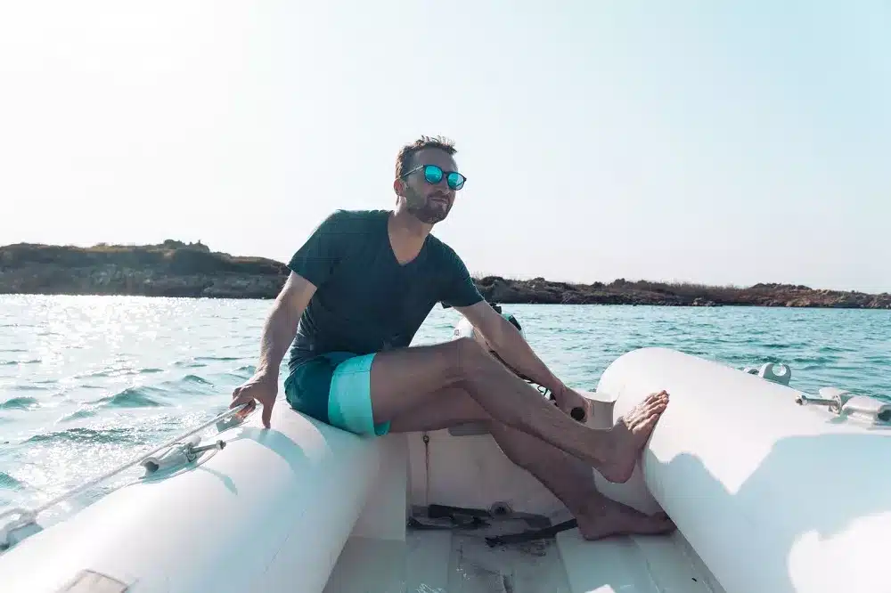 Ein Mann mit Sonnenbrille sitzt entspannt auf der Kante eines Katamarans und genießt einen sonnigen Tag auf dem Wasser nahe einer felsigen Küste.
