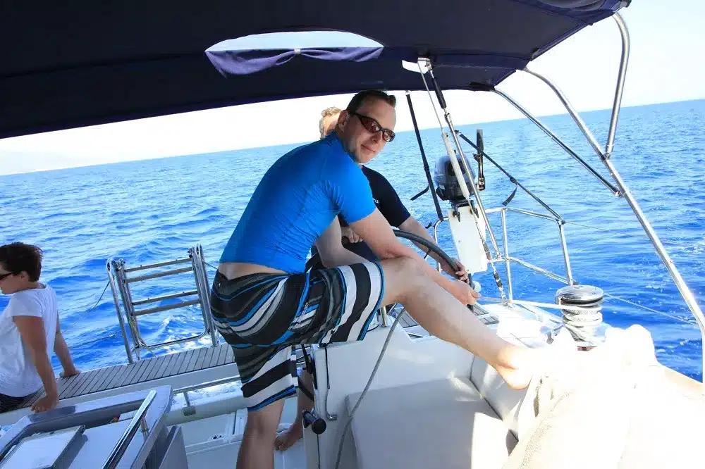 Ein Mann in einem blauen Hemd und gestreiften Shorts lächelt, während er an einem sonnigen Tag vom Steuer eines Katamarans steigt, mit klarem blauen Himmel und dem Meer im Hintergrund.