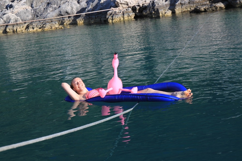 Eine Person entspannt sich in einem blauen Neoprenanzug auf einem aufblasbaren rosa Flamingo im klaren Wasser, im Hintergrund sind felsige Klippen und ein Katamaran zu sehen.