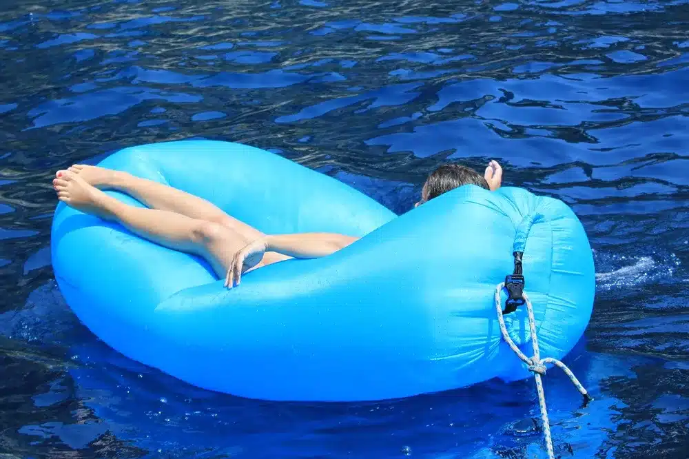 Eine Person entspannt sich auf einem blauen aufblasbaren Ring, der in dunkelblauem Wasser schwimmt. Nur ihre Beine und ihr Rücken sind zu sehen, da sie mit dem Gesicht nach unten liegt. Ein Seil, das mit dem Ring verbunden ist, erstreckt sich seitlich und ist an einem Katamaran befestigt.