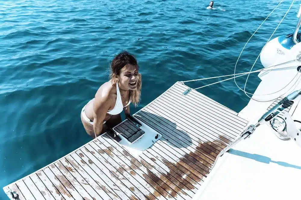 Eine Frau im weißen Badeanzug lacht fröhlich, während sie bei einem Segeltörn vom blauen Ozean aus auf das Deck eines Segelboots klettert, während eine andere Person daneben schwimmt.