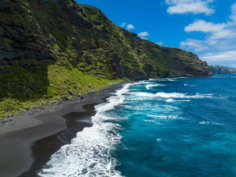 Luftaufnahme einer atemberaubenden Küste mit schwarzem Sandstrand, türkisfarbenem Wasser und üppig grünen Klippen unter einem klaren blauen Himmel, übersät mit Segelyachten.