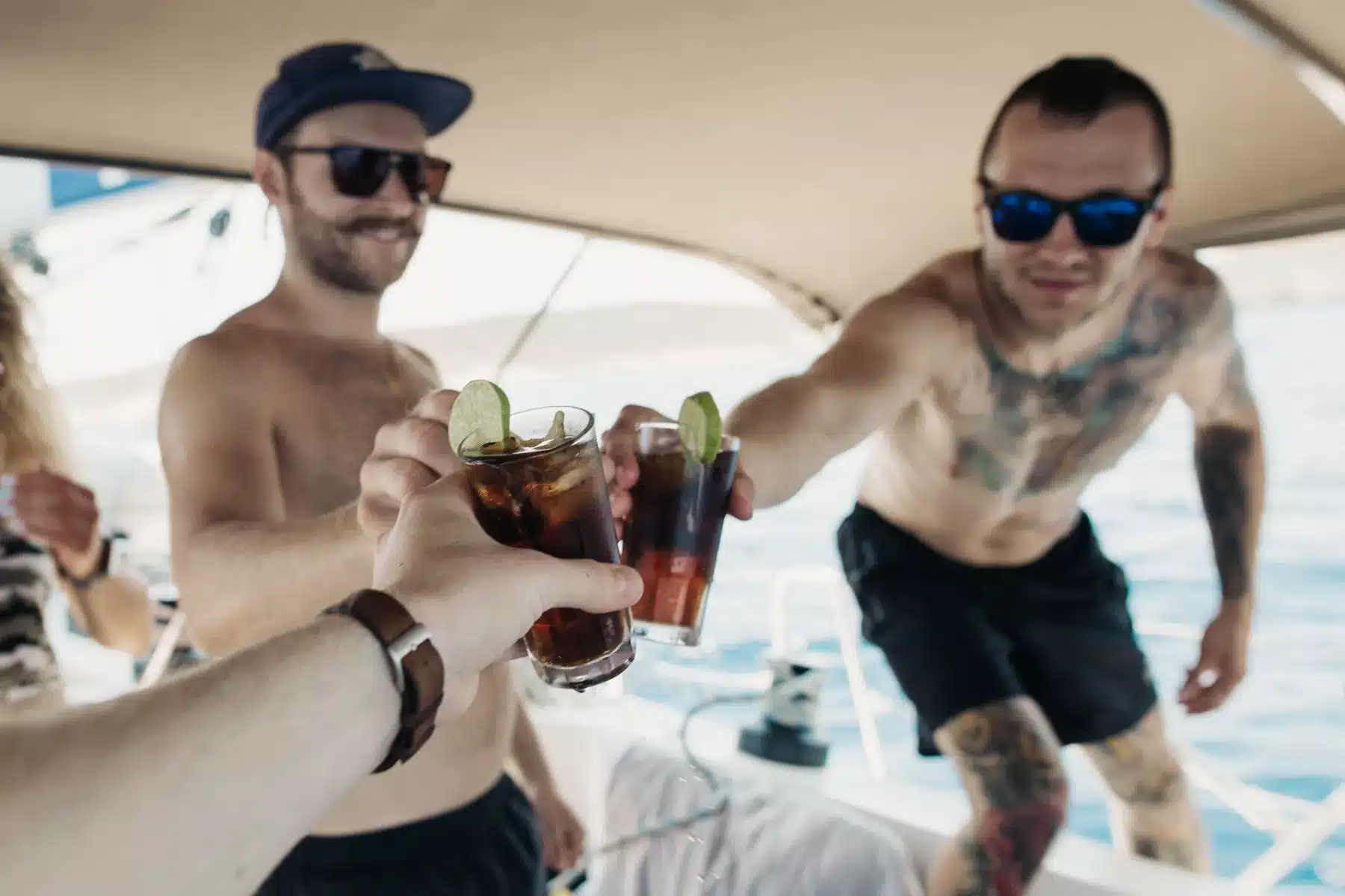 Drei Freunde stoßen auf einer Segelyacht mit eisgekühlten Getränken an. Im Vordergrund ist eine Hand zu sehen, die ein Getränk hält. Zwei von ihnen lächeln und sind leger gekleidet in einer sonnigen Umgebung.