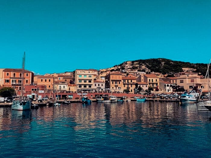Malerischer Küstenort mit farbenfrohen Gebäuden entlang der Uferpromenade. Boote, darunter ein eleganter Katamaran, liegen im ruhigen Hafen vor Anker und spiegeln sich im türkisfarbenen Wasser. Ein mit Grün bedeckter Hügel bietet eine Kulisse unter einem klaren, strahlend blauen Himmel, perfekt für ein Segelurlaubsabenteuer.