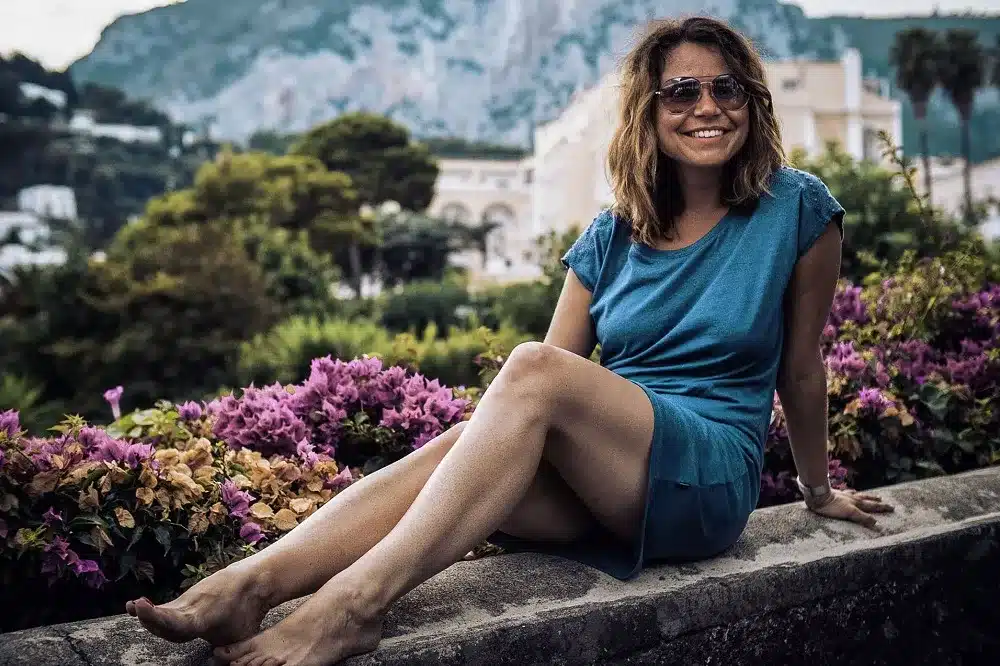 Eine Frau in einem blauen Kleid sitzt lächelnd auf einer Steinmauer, mit üppigem Grün und Bergen im Hintergrund, und träumt von ihrer nächsten Segelreise.