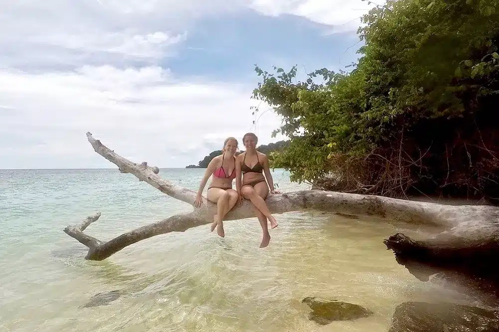 Zwei Personen sitzen auf einem großen Ast, der über das klare Wasser an einem Strand ragt, mit üppigem Grün und einer Segelyacht im Hintergrund.