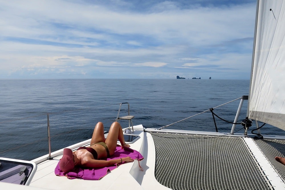 Eine Person im Bikini sonnt sich während eines Segeltörns auf dem Deck einer Yacht, mit einem klaren blauen Himmel über ihr und ruhiger See im Hintergrund. Badeurlaub auf dem Segelboot: Die schönsten Buchten entdecken