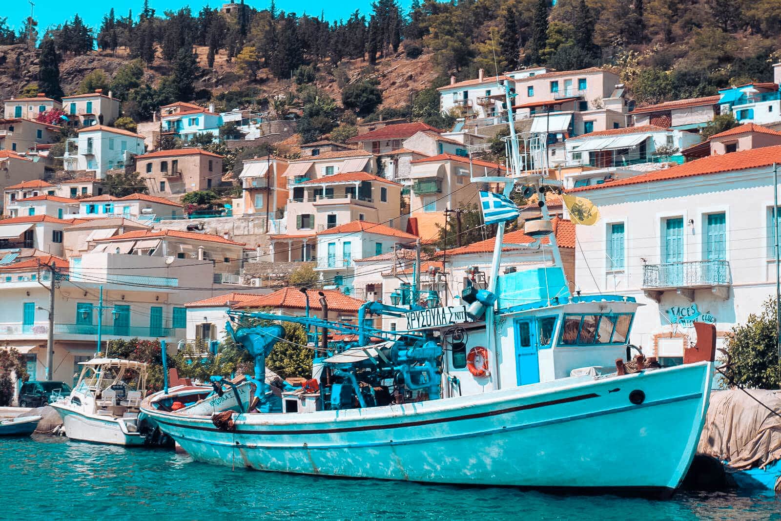 A picturesque coastal village with white and pastel-colored buildings on a hillside. In the foreground, a fishing boat with blue and white accents and fishing gear is docked. The sea is calm—perfect for a sailing trip—and in the background, a lush, tree-covered hill rises.