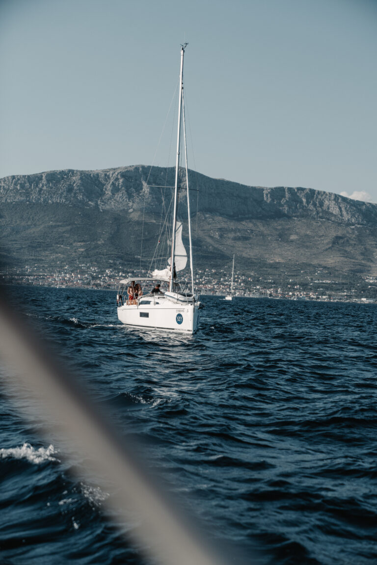 Ein Katamaran mit mehreren Personen an Bord schwimmt auf dem Meer, im Hintergrund ist eine Berglandschaft unter einem klaren Himmel zu sehen.