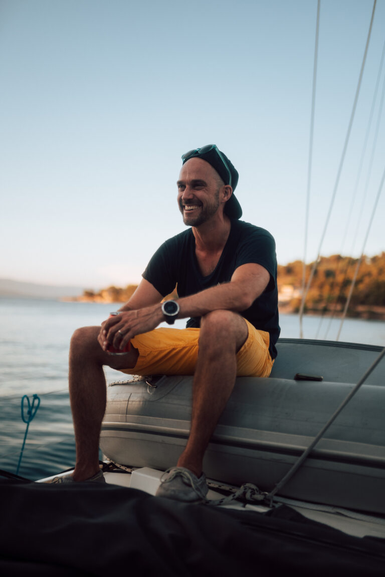 Ein gut gelaunter Mann sitzt mit Mütze, T-Shirt und gelben Shorts auf dem Deck einer Segelyacht und genießt den Sonnenuntergang am Wasser.