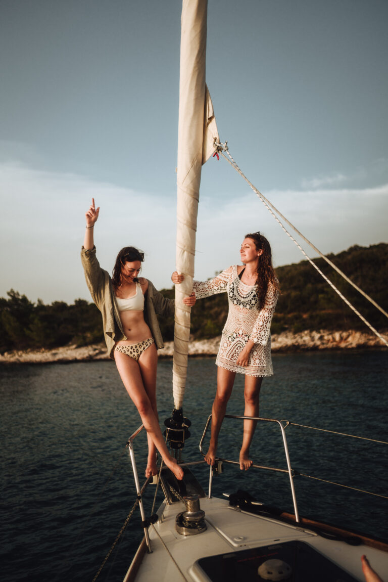 Zwei Frauen posieren fröhlich auf dem Deck eines Katamarans, eine streckt ihre Hand in die Luft, umgeben von einer ruhigen, von Bäumen gesäumten Küste.