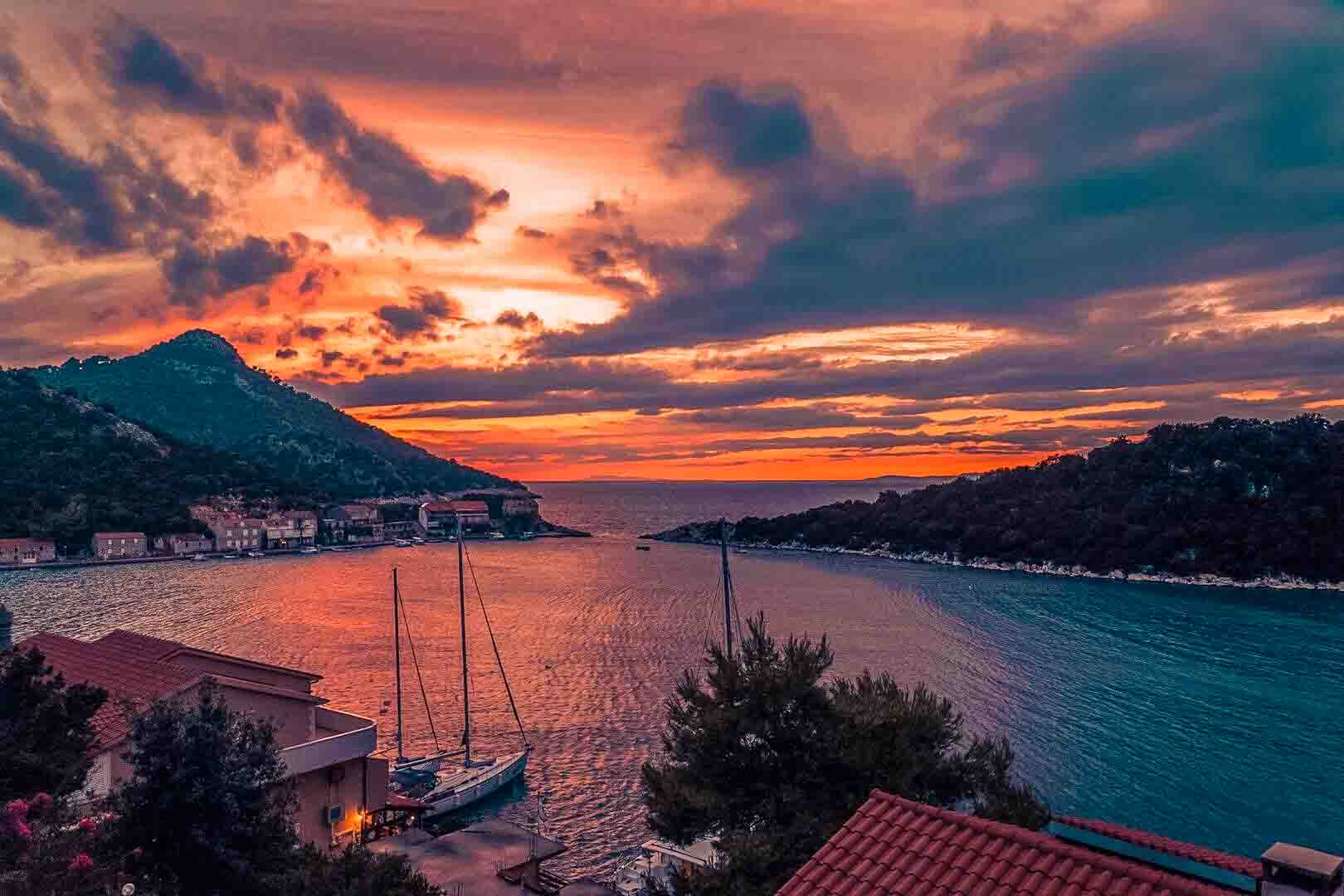 Ein malerischer Sonnenuntergang über einer Küstenstadt mit Segelbooten und einem eleganten Katamaran, der am Ufer angedockt ist. Der Himmel ist eine Mischung aus leuchtenden Orange-, Rosa- und Lilatönen, die sich im ruhigen Wasser spiegeln. Hügel und Dächer rahmen die ruhige Szene ein und schaffen eine friedliche Atmosphäre.