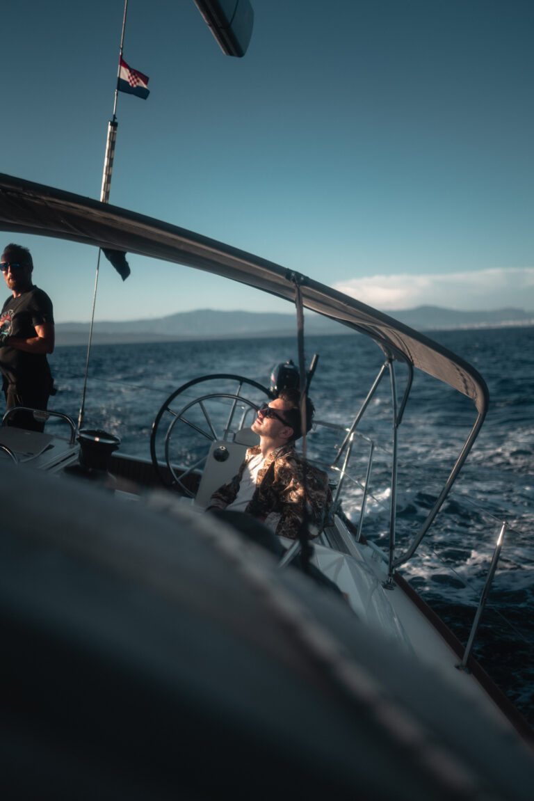 Eine Frau steuert eine Segelyacht und blickt auf die Segel, im Hintergrund das Meer und den blauen Himmel. Eine andere Person steht in der Nähe und auf dem Boot weht eine amerikanische Flagge.