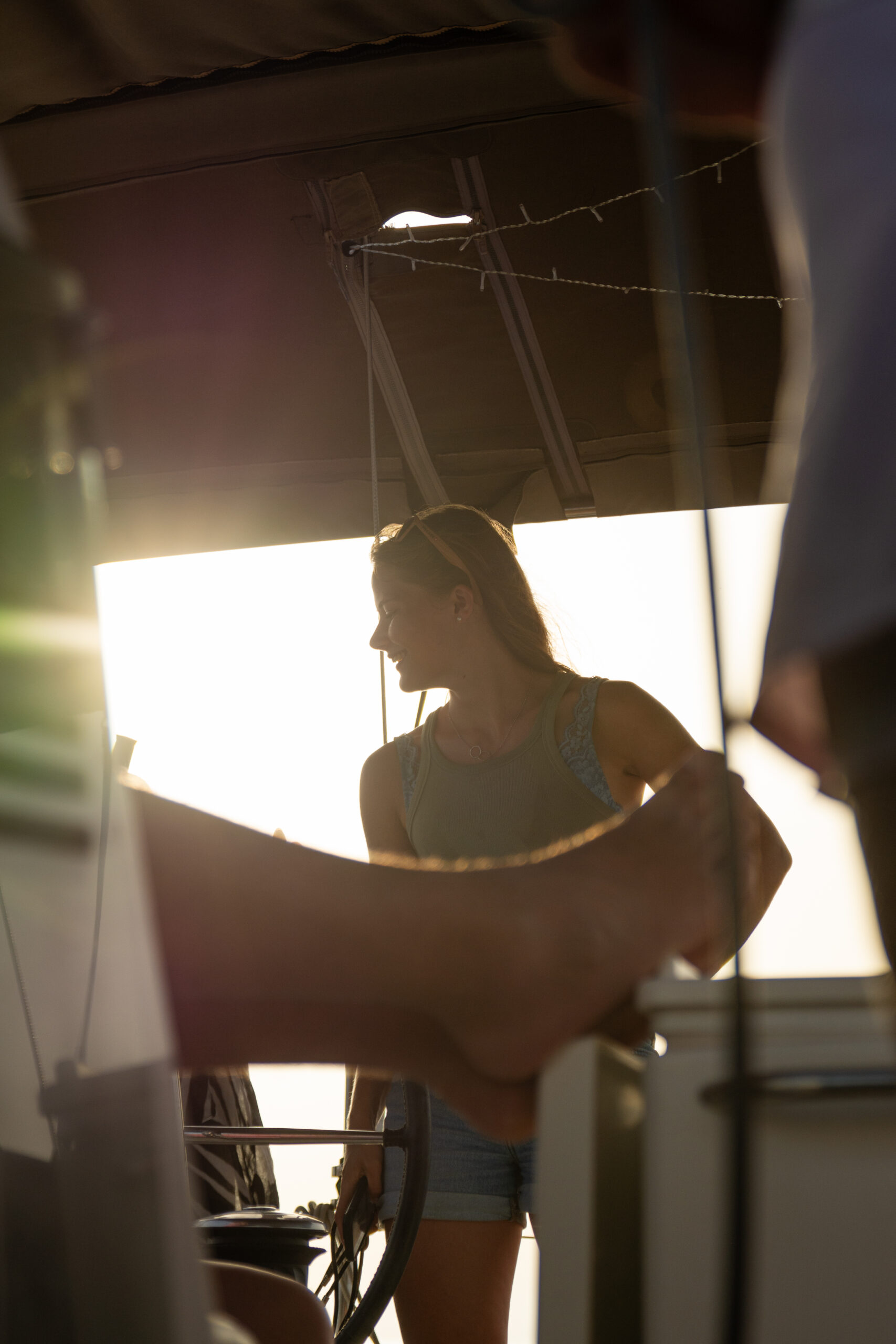 Frau steht auf einer Segelyacht, im Gegenlicht der Sonne, und unterhält sich mit einer Person, deren Arm im Vordergrund zu sehen ist. Die Umgebung ist ruhig und strahlt warmes Licht aus.