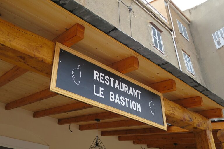 Ein Holzschild für „Restaurant Le Bastion“, das unter einem Holzbalken vor einer sonnigen, beigen Gebäudefassade hängt. Das Schild zeigt dekorative Umrisse einer Kochmütze und eines Katamarans.