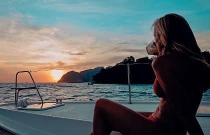 A woman in a red dress sits on a catamaran, gazing at the sunset over the sea, with the silhouette of an island in the background.