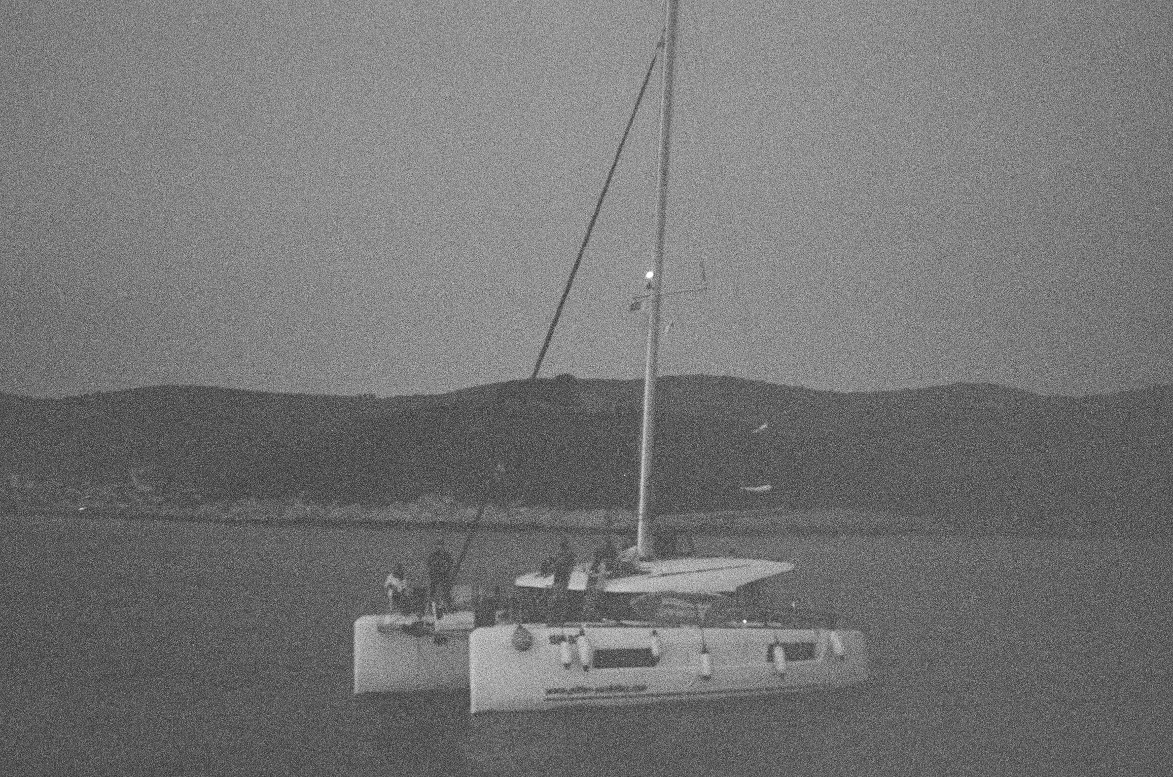 Ein Schwarzweißfoto eines Katamarans, der in der Abenddämmerung in der Nähe einer hügeligen Küste segelt, wobei das Wasser nur minimales Licht reflektiert.
