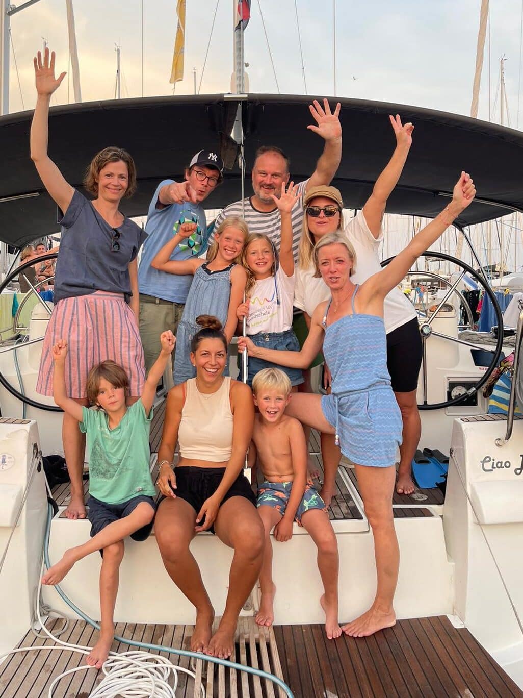 Eine Gruppe von zehn Personen, darunter Erwachsene und Kinder, posiert gemeinsam auf einem Boot, lächelt und hebt die Hände. Sie scheinen einen sonnigen Tag im Yachthafen zu genießen, im Hintergrund sind Segelboote zu sehen, bereit für ihr Segelreise-Abenteuer.