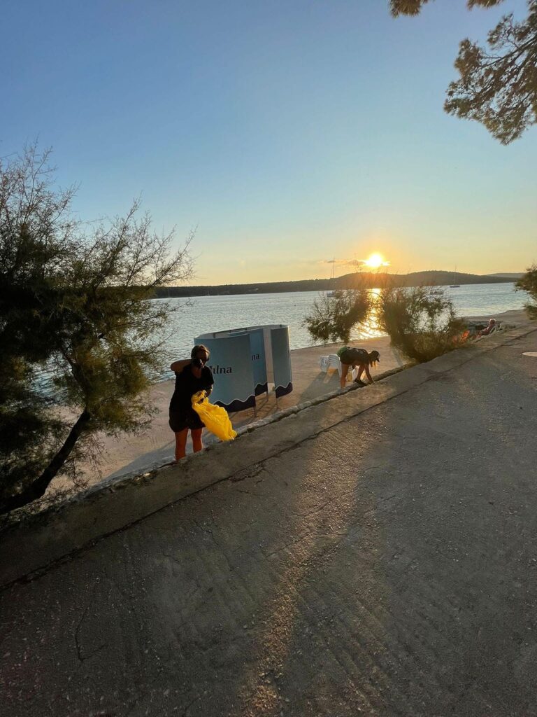 Sunset over a calm lake with people walking along the shore, some carrying items. Trees partially shade the path, and a bright sun hovers above the horizon. A catamaran sails gently across the water.