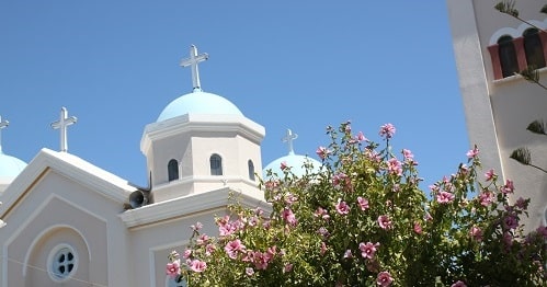 Kirche mit blauer Kuppel und weißen Wänden unter klarem Himmel, flankiert von einem Glockenturm und umgeben von rosa blühenden Blumen, mit Blick auf eine Segelyacht.